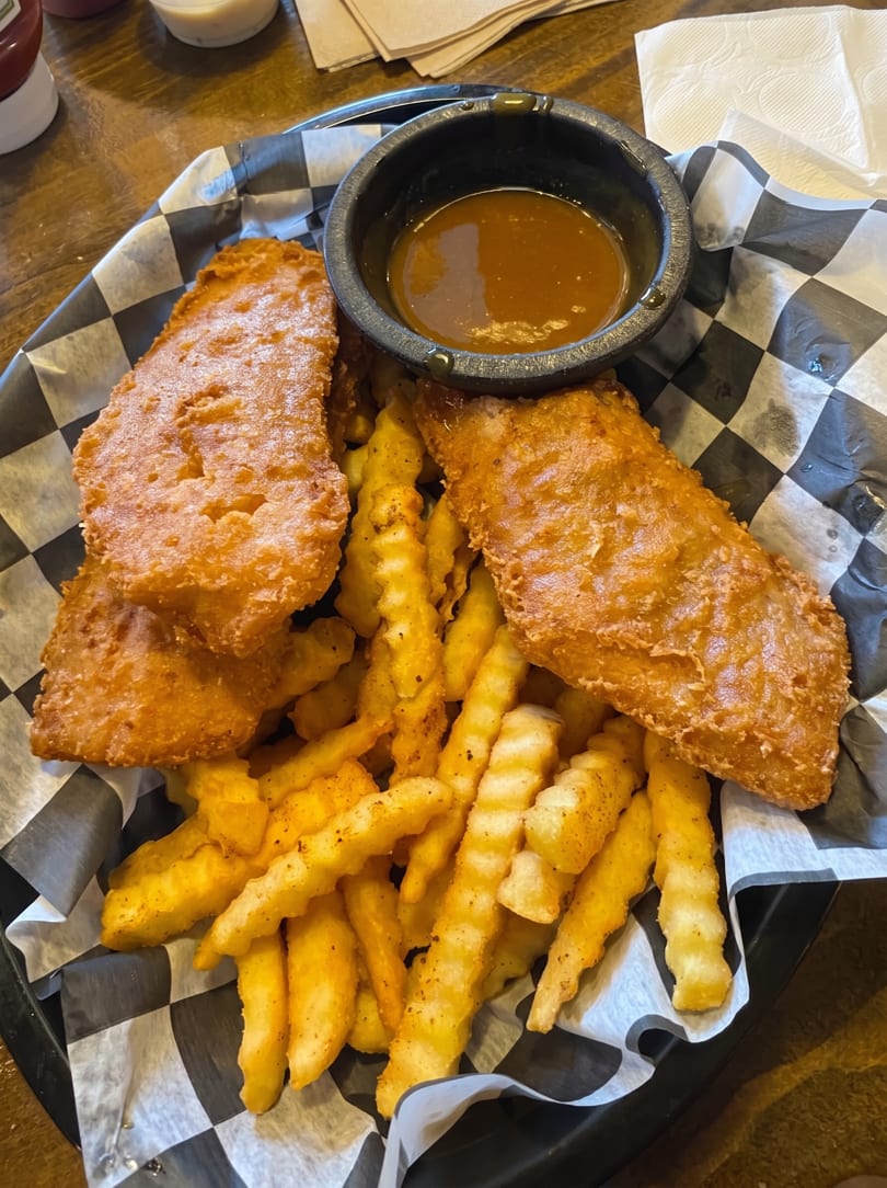 Golden-brown fish and chips with a side of brown dipping sauce in a black basket.